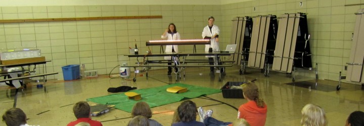 two people teaching children using a horizontal tube with flames on top